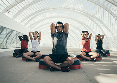 People meditating on step platforms under a modern, arched structure. They wear athletic gear in black, red, and white, creating a calm scene.