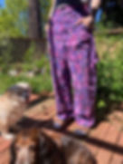 a shot highlighting a pair of bright purple floral-patterned pants, in a wide-leg climbing pants style, worn with sandals and socks on a deck with an overgrown garden in the background. Two dogs are also chilling on the deck