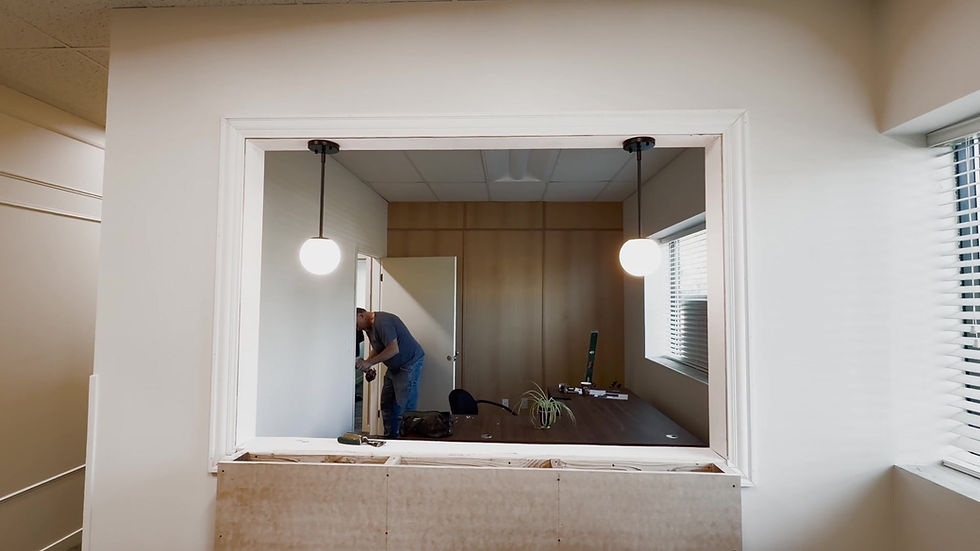 Work underway behind a wide interior opening, with two globe pendants, a wood-tone paneled wall and an unfinished lower ledge.