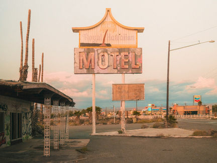 L'insegna logora del Royal Hawaiian Motel di Arne in uno scenario lungo una strada nel deserto al crepuscolo, con palme, linee elettriche e una stazione di servizio Chevron.