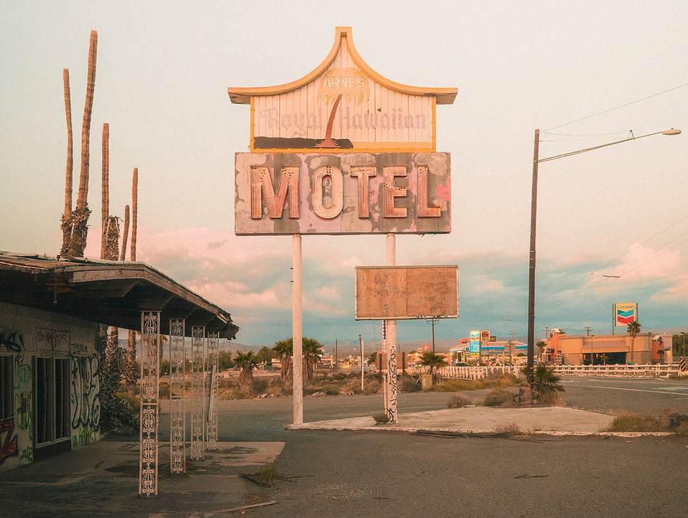 L'insegna logora del Royal Hawaiian Motel di Arne in uno scenario lungo una strada nel deserto al crepuscolo, con palme, linee elettriche e una stazione di servizio Chevron.