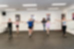 Group of people in a dance studio practicing with arms outstretched. Mirrors and framed posters on walls, light-colored flooring.