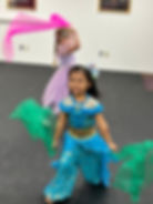 Two children in costumes dance joyfully with colored scarves in an indoor setting. One wears blue and gold, the other lavender, on a black floor.