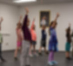 Children in colorful outfits stand at Tamraloo Studios in Lincoln, CA with arms raised as they learn how to dance. A framed picture hangs on the wall, creating a focused, playful mood.