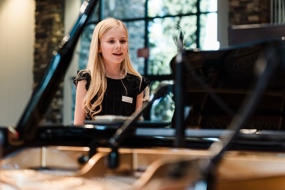 Girl in a black dress sings at a grand piano in a bright room, focused and poised.