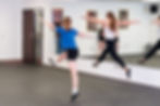 Two dancers leap in sync in a studio with mirrored walls. One wears blue and black, the other white and black. Others observe in the background.