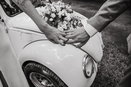 Twee handen op motorkap met bloemen op witte Kever – detail van een klassieke trouwdag in Zeeland