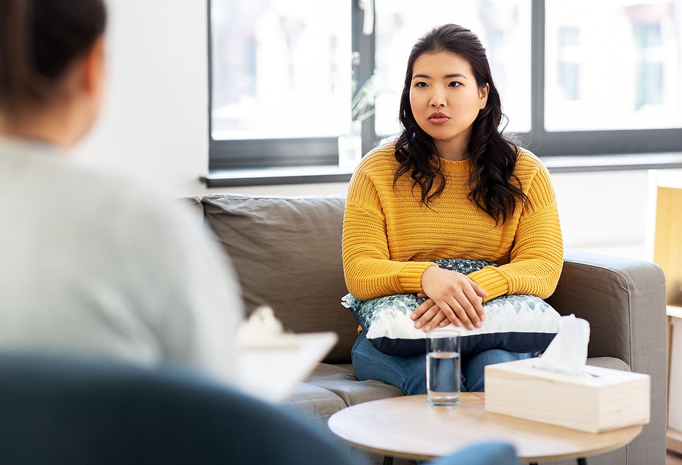 Woman in yellow sweater holding pillow, sitting on gray couch, appears engaged in conversation. Indoor setting with bright window.