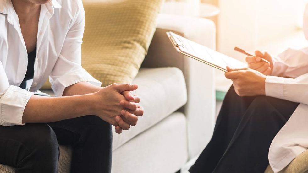 Two people sit on a sofa during a counseling session. One holds a clipboard, the other with clasped hands. Soft lighting creates a calm mood.