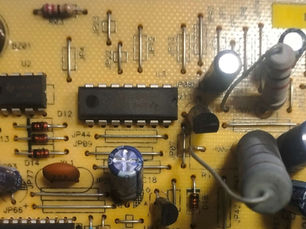 Gambar: Close-up rangkaian PCB menampilkan resistor warna coklat, IC hitam, kapasitor elektrolit biru, dan komponen elektronika lainnya pada board kuning.