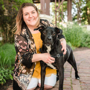 Alana smiling, crouching beside a black dog on a brick path. Wearing a floral kimono and yellow top. Lush greenery in the background.