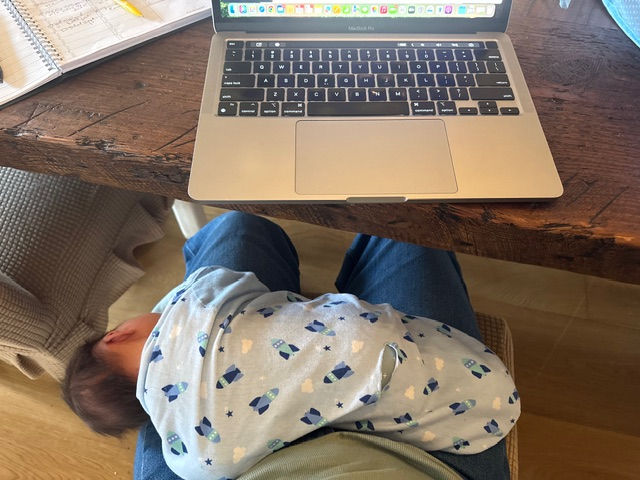 A woman working on computer with a napping baby on her lap.