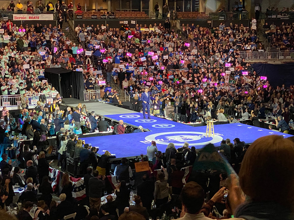 Pete Buttigieg walks out to the stage at the McIntyre-Shaheen Dinner in front of the section of Bernie Sanders supporters with lighted signs.
