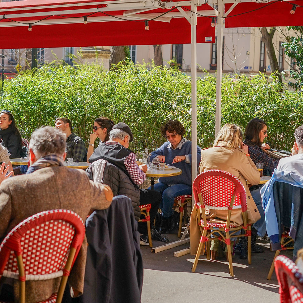 Terrasse de café en plein air sous auvents rouges, clients assis à des tables, discutant et mangeant, ambiance conviviale au soleil.