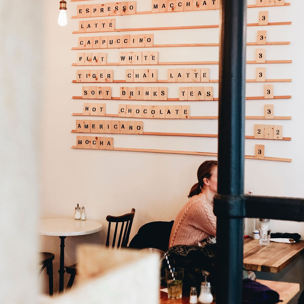 Intérieur de café avec menu en bois (espresso, latte, cappuccino) et une femme assise à table, ambiance douce.