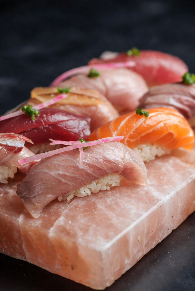Assorted nigiri sushi with tuna and salmon on a Selrox pink salt block. Vibrant colors, garnished with green herbs and pink strips. Dark background.