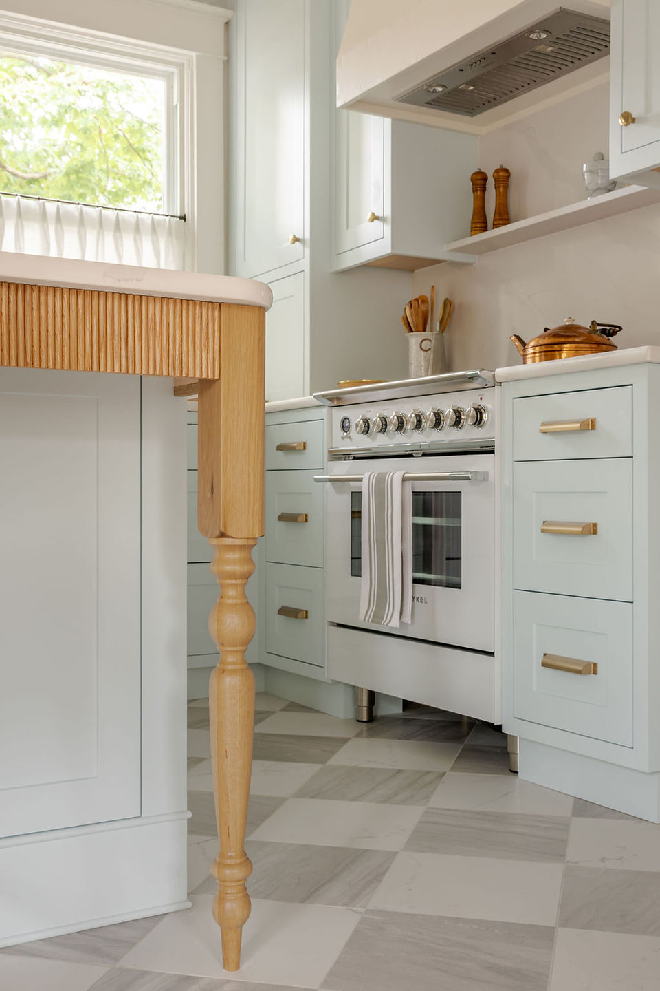Reeded oak island leg with a marble countertop beside a white range and sage green cabinetry on checkerboard marble floors.