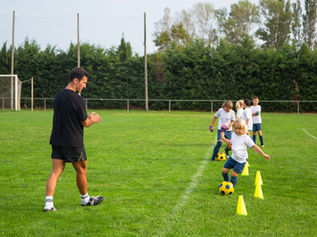 Kids in white shirts and blue shorts dribble yellow balls around cones on a grassy field, as a coach in black watches and claps.