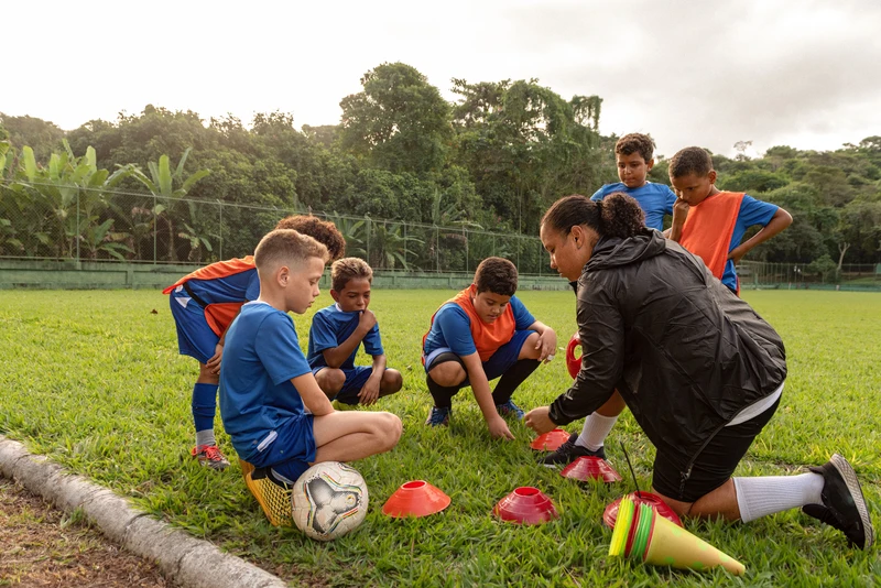 A football coach instructing children while kneeling on a football field surrounded by coaching equipment such as ball and cones. 