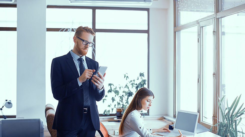 Businessman in suit uses a smartphone beside a woman typing on a laptop in a bright modern office.