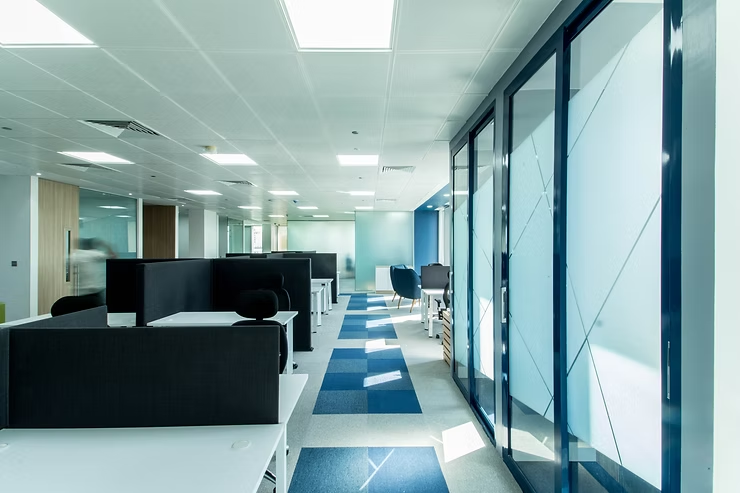 Bright open-plan office at Verod Capital with blue-accented carpet, ergonomic desks, and glass-partitioned meeting rooms in a corporate workspace.