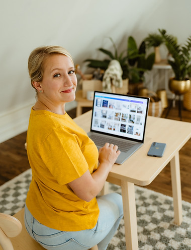 A woman smiling, wearing a yellow shirt typing on a laptop and looking back at the camera