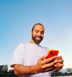 Ein lächelnder Mann im weißen T-Shirt schaut draußen auf sein orangefarbenes Smartphone.