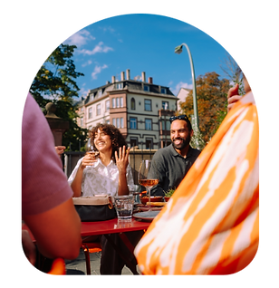 Eine Gruppe unterhält sich lachend an einem Tisch auf einer sonnigen Café-Terrasse in der Stadt.