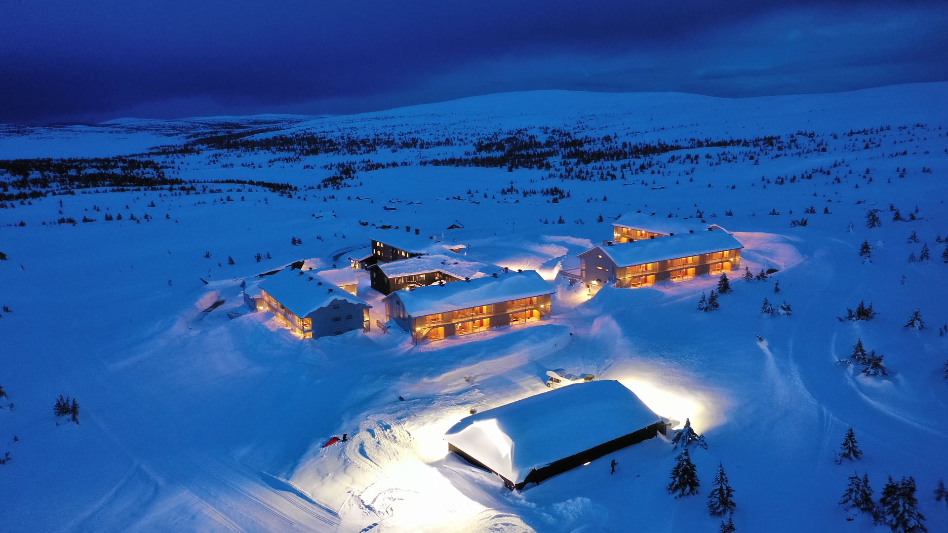 Opplyste bygninger omgitt av snø i blått kveldslys, sett ovenfra.