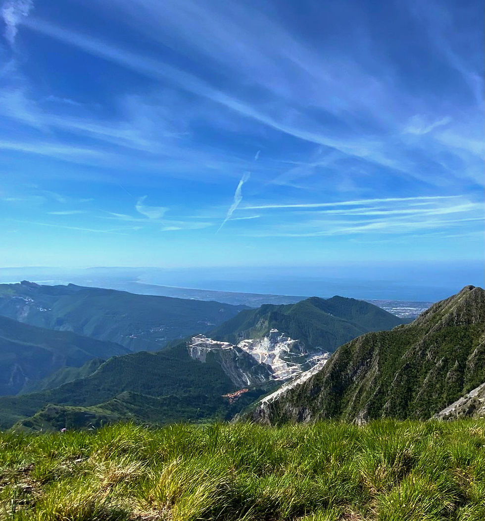 Blick auf grüne Berge und einen Marmor Steinbruch in Carrara unter blauem Himmel mit dünnen Wolkenstreifen. Im Vordergrund saftiges Gras. In der ferne sieht man Meer der versilia