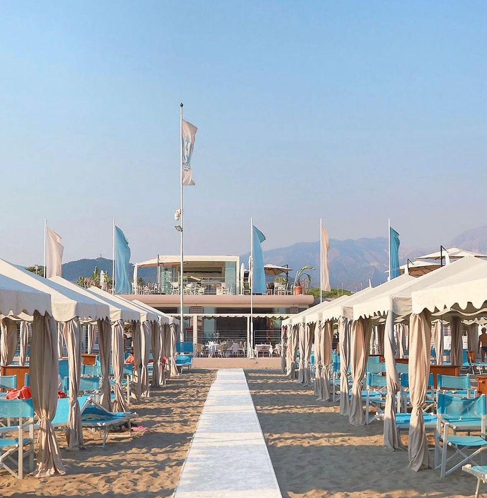 Weiße Zelte säumen einen steinernen Weg zu einem Strandpavillon. Blau-weiße Flaggen wehen, Berge der apuanischen Alpen im Hintergrund unter blauem Himmel.