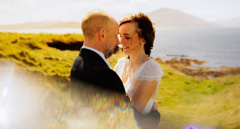 Bride and groom embracing outdoors near a grassy cliffside with the ocean and distant mountains in the background, both smiling with foreheads touching in a sunlit, romantic moment.
