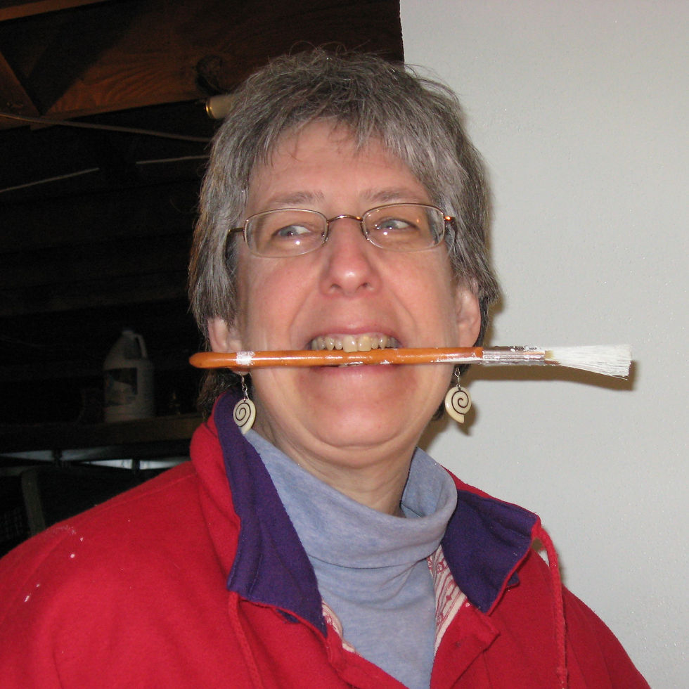 Barb Bickford holding a paintbrush in her teeth and looking off to the side mischievously. Photo credit: Aralee D.