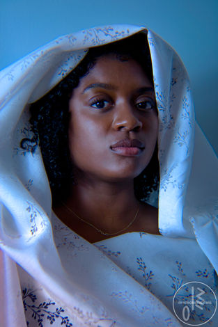 2024 portrait photograph by Lewis Derogene, artistically known as PhenomenaLewis of model Lisa Jean Felix. This photograph, "The Girl With the Soft Gaze," was taken with the intent of representing a Black woman in a soft visual as a form of resilience against stereotypical tropes. Photographer: Lewis Derogene. Model: Lisa Jean Felix. Photo description: A Black woman veiled in a white floral lace with soft blue lighting and a pink lighting in the corner with part of her curly hair poking out of the veil as she looks directly at the camera.
