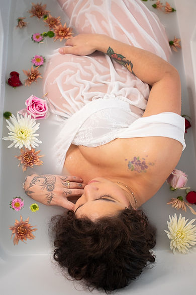 A pregnant woman in a sheer white dress lays in a milk bath surrounded by beautiful flowers. She is relaxed and content.