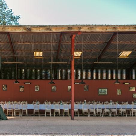 Covered outdoor dining area with long tables, white tablecloths, and empty chairs. Green curtains and paintings on red walls. Serene and ready.