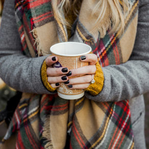 Person in gray coat, plaid scarf holding a coffee cup with black nail polish and ring. Warm street setting, festive background. Cozy mood.