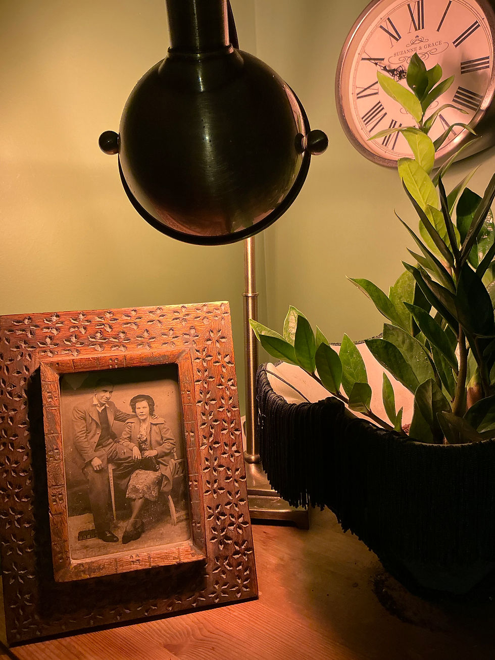 Warm indoor scene with a vintage framed photo of a couple on a wooden table, lit by a lamp, with a plant and a clock in the background
