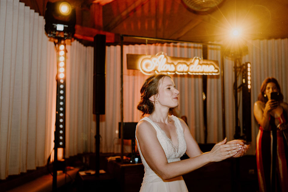 Photo de mariage signée Julien Loize, photographe professionnel à Soissons : la mariée applaudit joyeusement sur la piste de danse, avec un panneau lumineux 'Alors on danse' en arrière-plan, lors d'une soirée en Picardie.