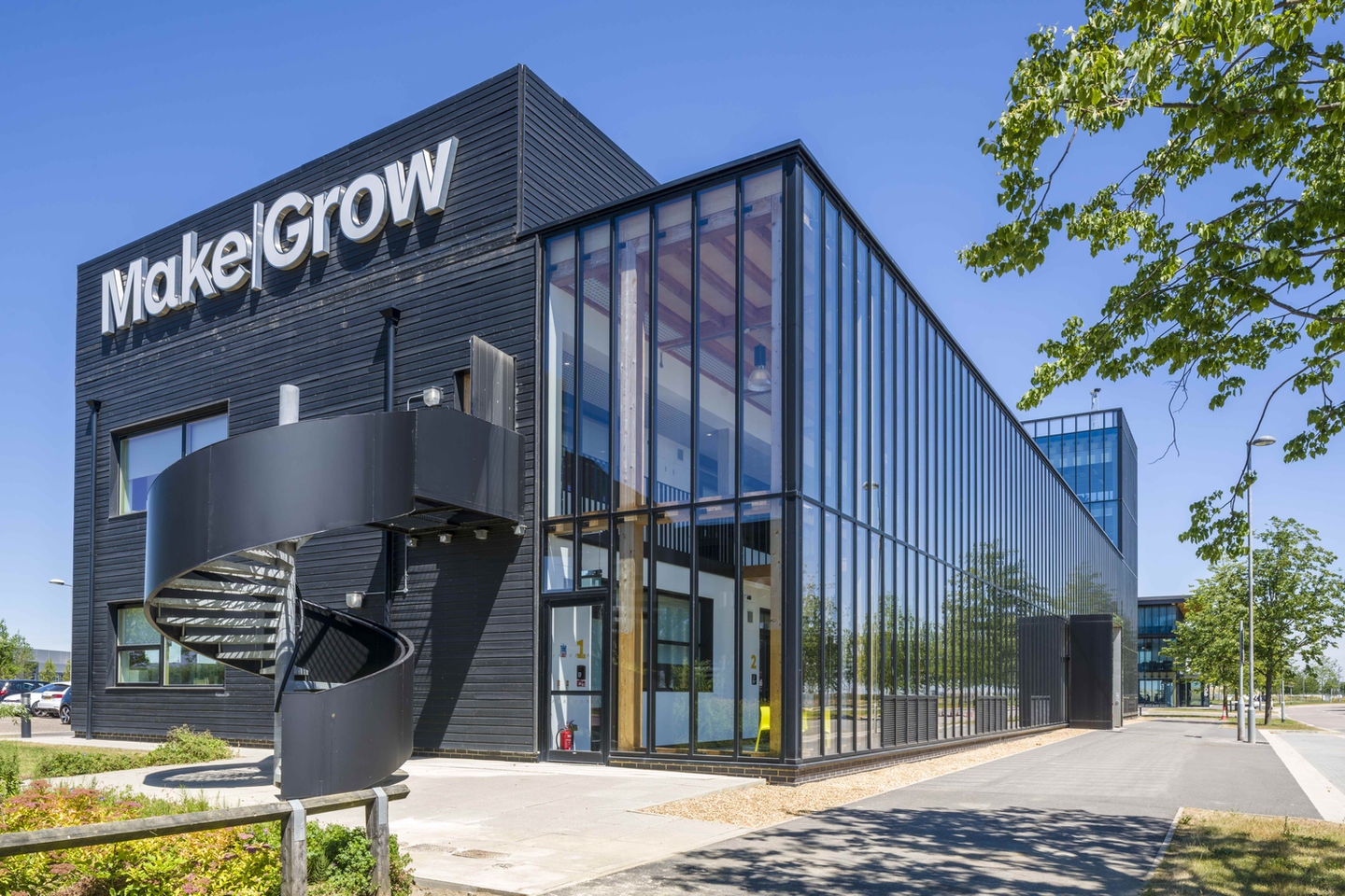 A modern sleek black building displaying "Make|Grow" on its exterior. There is extensive use of glass, providing a transparent view of the interior. A distinctive black spiral staircase is attached to the side of the building. The surroundings are well-maintained with a clear path, neatly trimmed plants, a few trees and a clear blue sky.