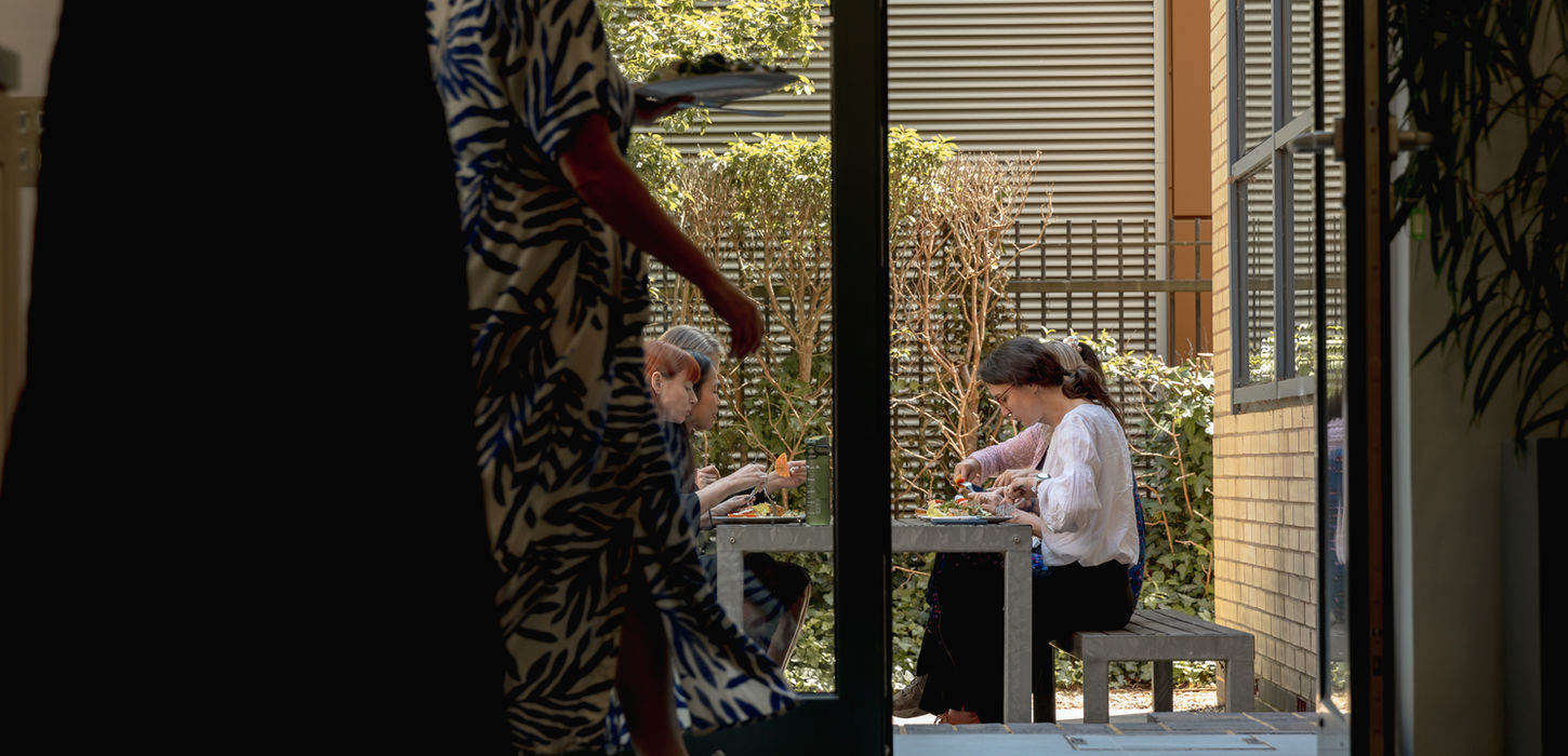 A group of people sitting at a table outdoors, enjoying a meal, as seen through a doorway. A person in a patterned dress walks by in the foreground, and green plants and modern architecture are visible in the background.