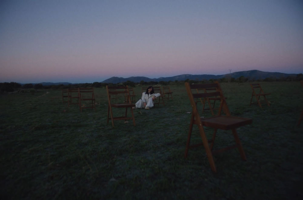 Lone person sits in a field with scattered chairs at dusk.