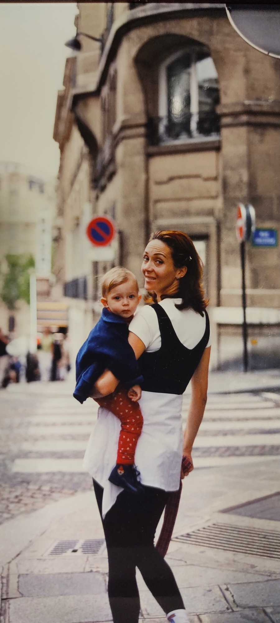A tall brunette woman stands on a street in Paris with her young daughter on her hip. She looks over her shoulder as her daughter looks into the camera. Taken in early 2000s, France.