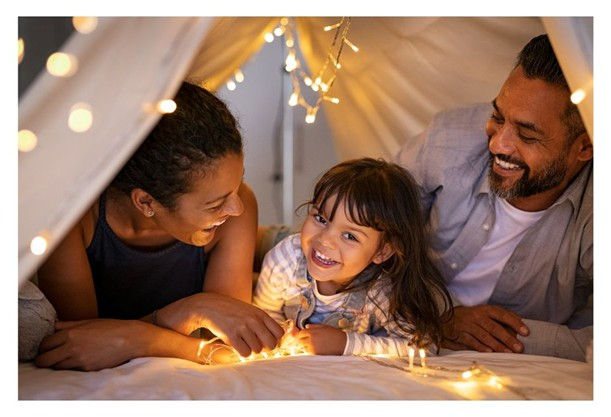 Family of three smiling under a cozy blanket fort, surrounded by warm fairy lights, creating a joyful and intimate atmosphere.