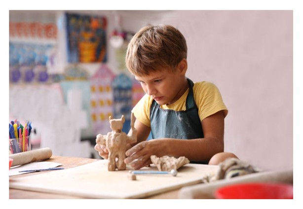 A child in a yellow shirt sculpts a clay figure on a table, focused amid colorful art supplies. Background shows vibrant artwork.
