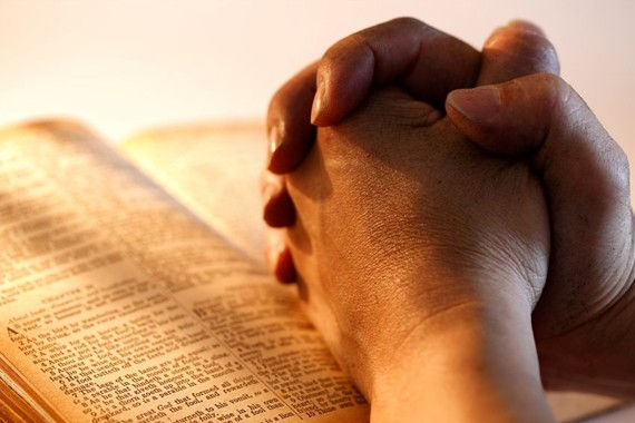 Hands clasped in prayer over an open Bible with visible text. Warm lighting creates a calm, reflective mood, highlighting the pages.