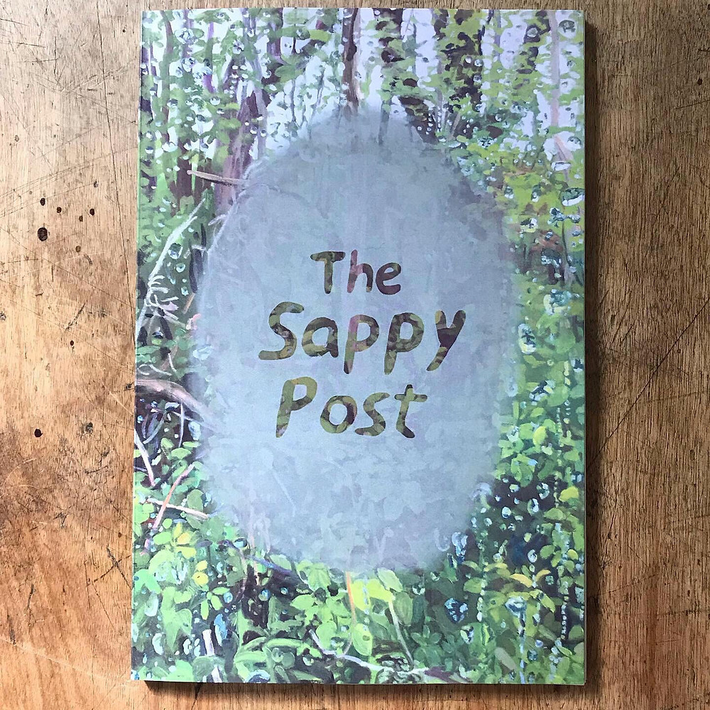 The Sappy Post on a wood table. The cover has a painting of a forest, with water droplets. The title looks like someone has breathed on glass and then written in with their finger.