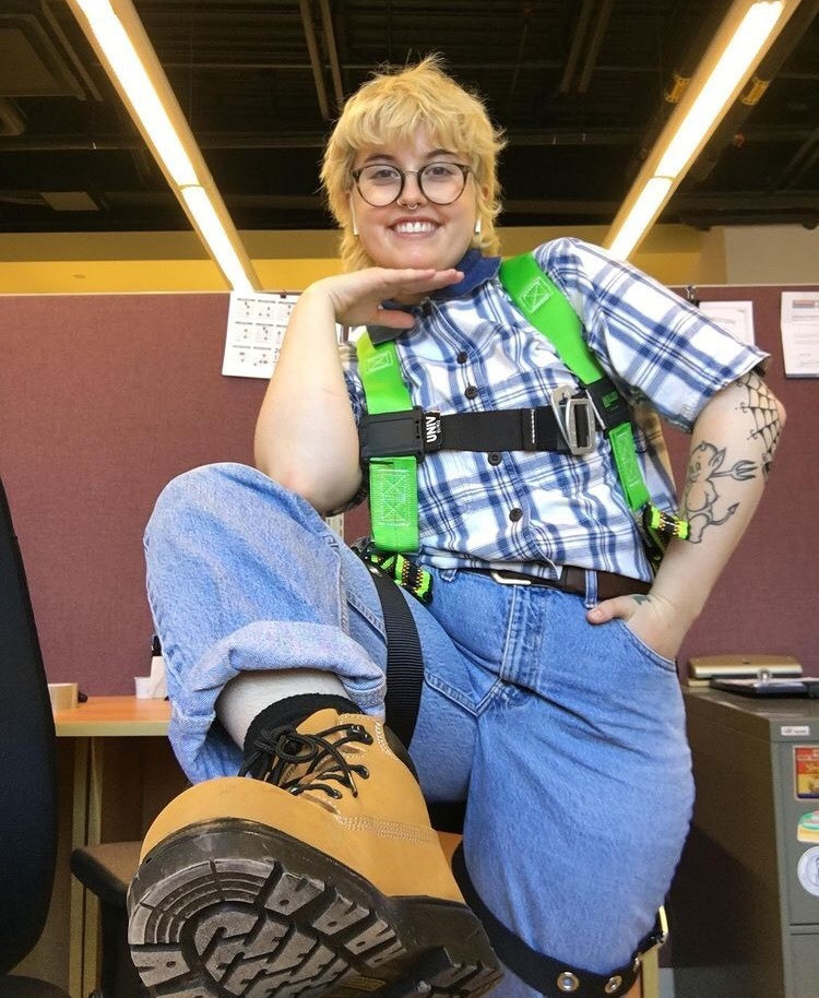 A photograph of a smiling person with one hand in their pocket, and one under their chin, with one foot up on a table in a dimly lit office. They are white, with short bleached hair, glasses, and tattoos, wearing a blue plaid shirt, jeans, a climbing harness, and brown hiking boots.