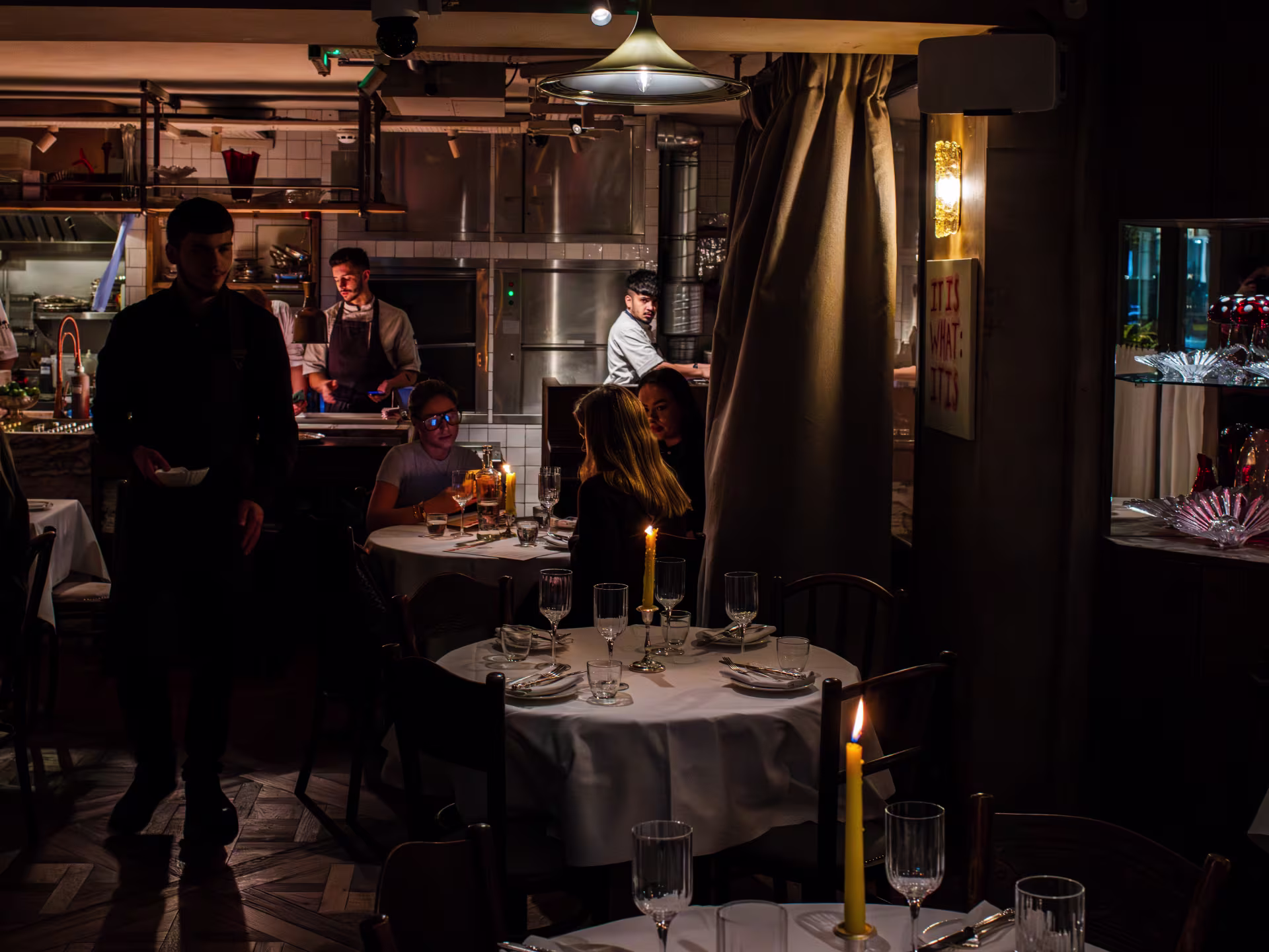 A dark intimate restaurant with candle lit tables and an open kitchen 