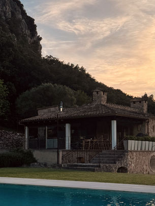 Stone house with tan brick and glass facade, lush green trees, and a rectangular pool in the foreground. Mountains and dusk sky in the background.